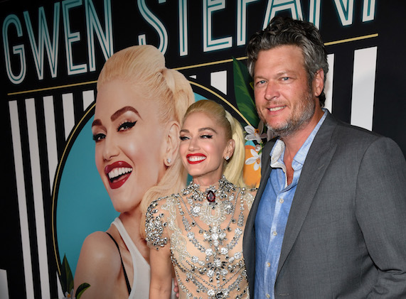 Gwen Stefani and Blake Shelton stand smiling on red carpet with photo of Gwen Stefani behind them