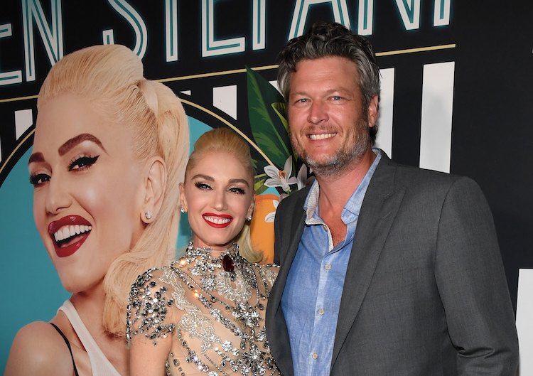 Blake Shelton and Gwen Stefani stand smiling with photo of Gwen behind them
