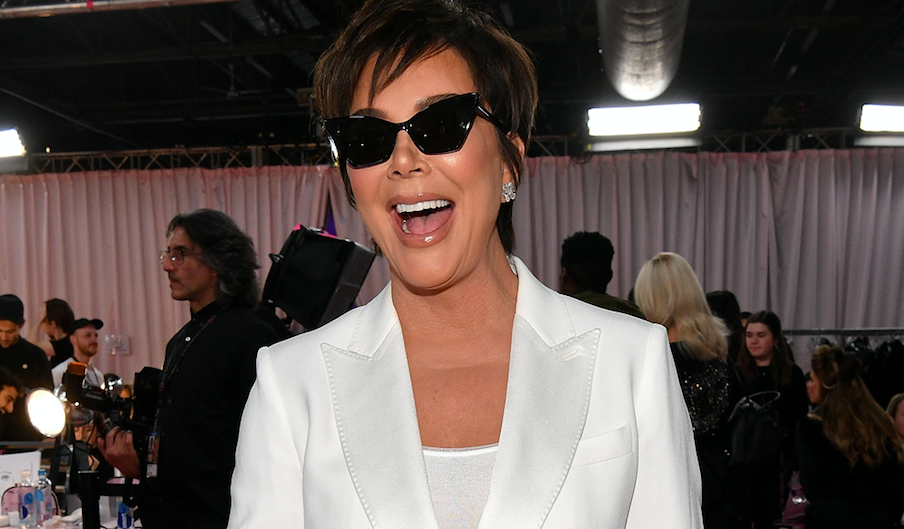 Kris Jenner smiling while wearing black sunglasses and a white pant suit