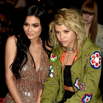 Kylie Jenner and Sofia Richie sitting together at New York Fashion Week in 2017, Kylie is wearing a sparkly gold dress and Sofia is wearing a green jacket with patches on it