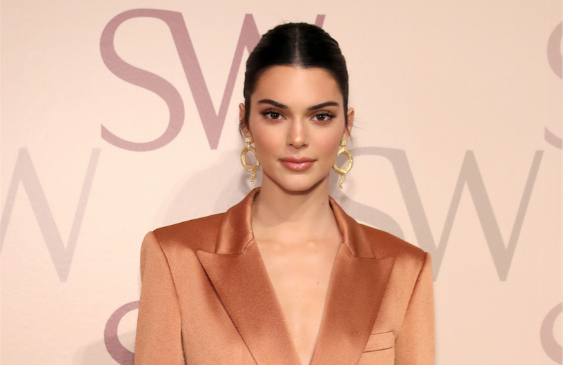Kendall Jenner posing wearing an orange jacket with her hair up and gold earrings
