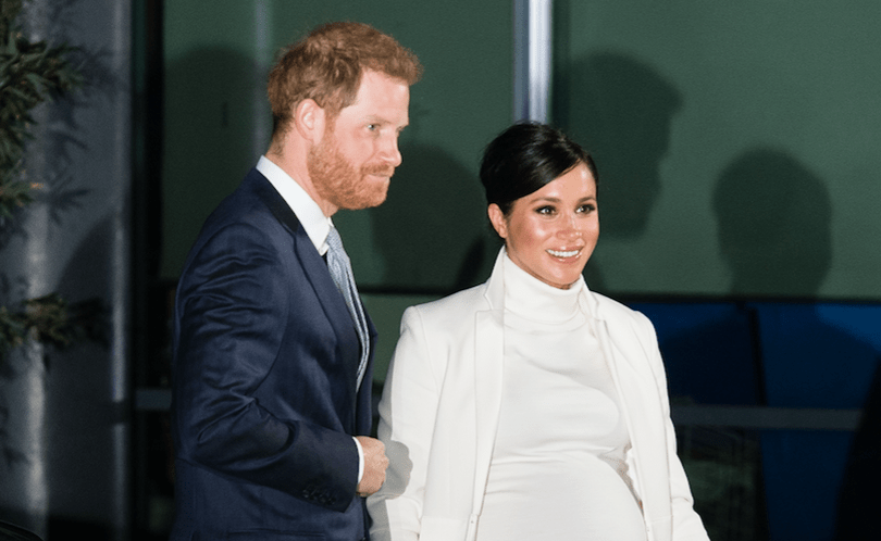 Prince Harry wearing a navy suit with his hand on Meghan Markle's back as she wears a white dress and jacket