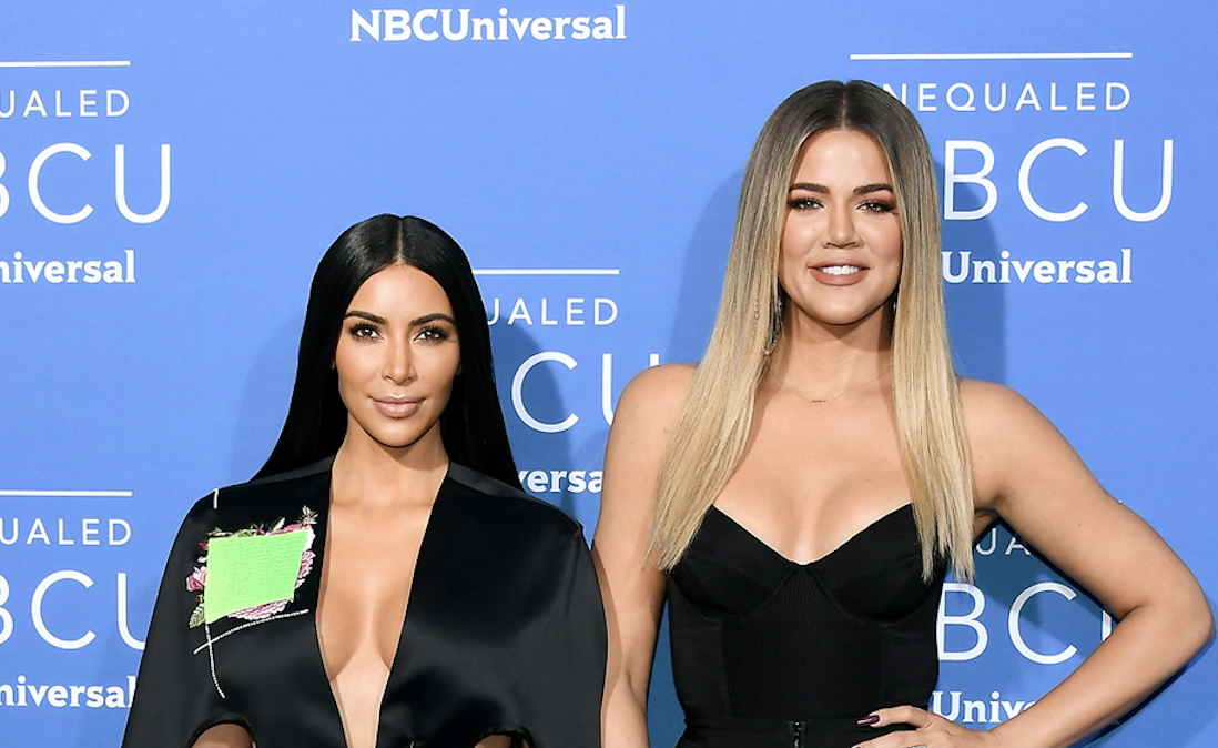 Kim Kardashian and Khloe Kardashian posing in front of a blue backdrop wearing all black.