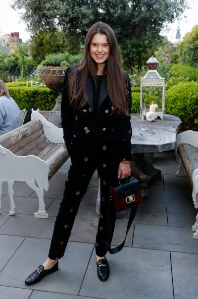 Model Sarah Ann Macklin Poses in a Black Suit With Roses All Over It While Smiling With Her Long Brown Hair Straightened 