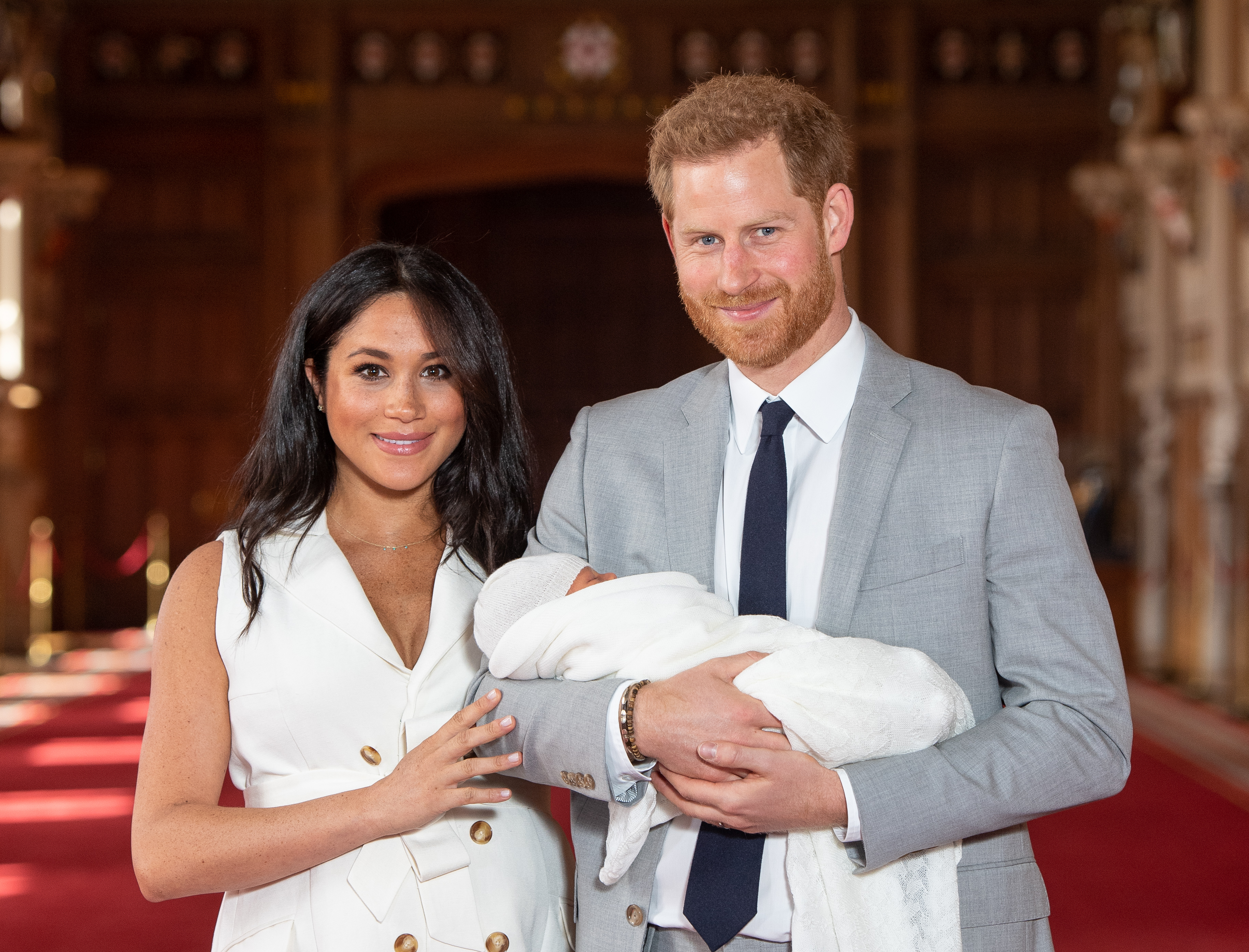 Meghan Markle Smiles in a White Trench Coat Style Dress Standing Next to Prince Harry in a Grey Suit and Navy Blue Tie They Are Holding Baby Archie Who Is Wrapped in White Swaddling Clothes