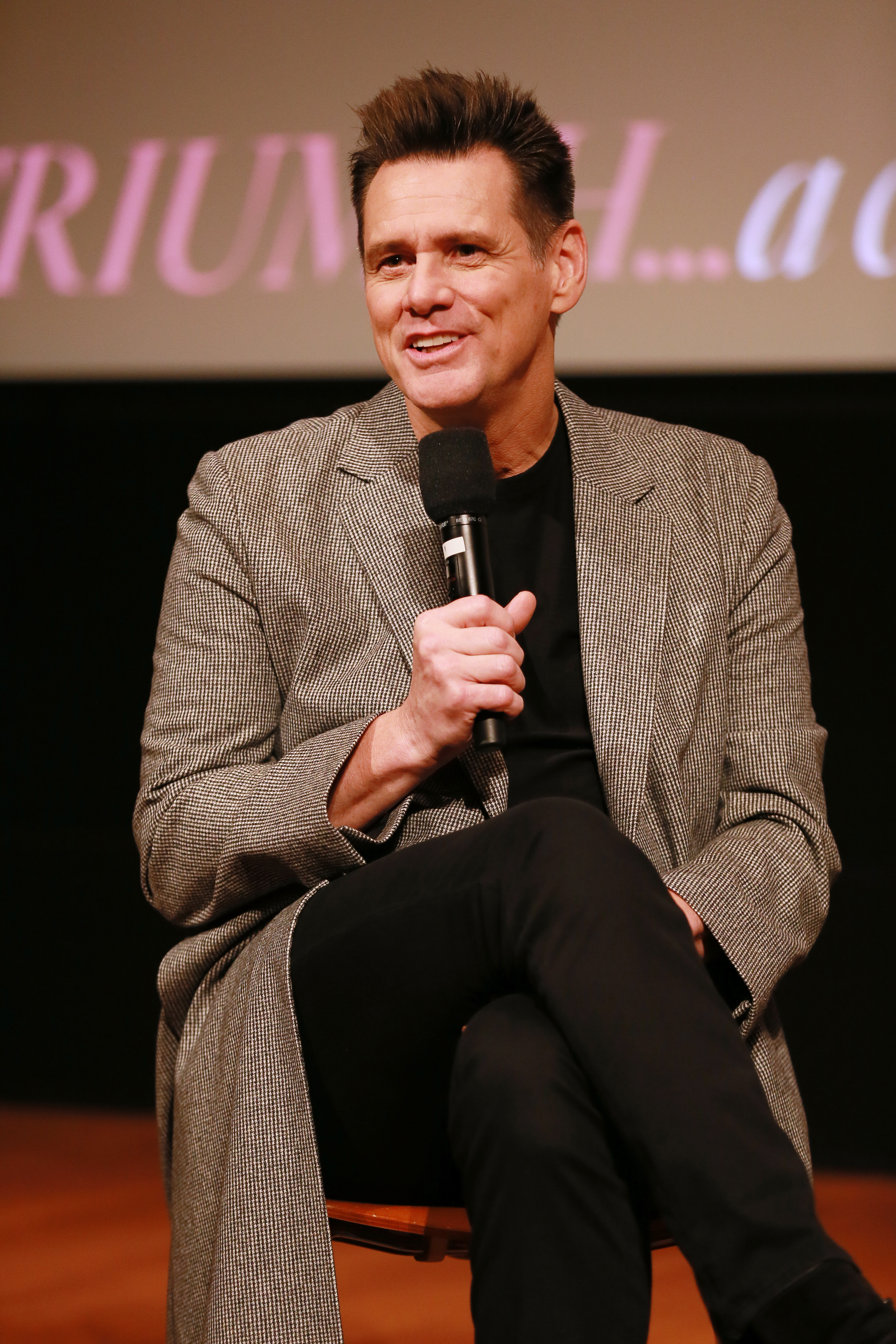 Jim Carrey Sitting in a Chair Holding a Microphone in a Black Outfit With a Long Tan Jacket