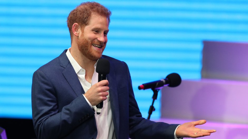 Prince Harry Shrugs and Smiles in Navy Blue Blazer With White Button Down Shirt While Holding a Microphone