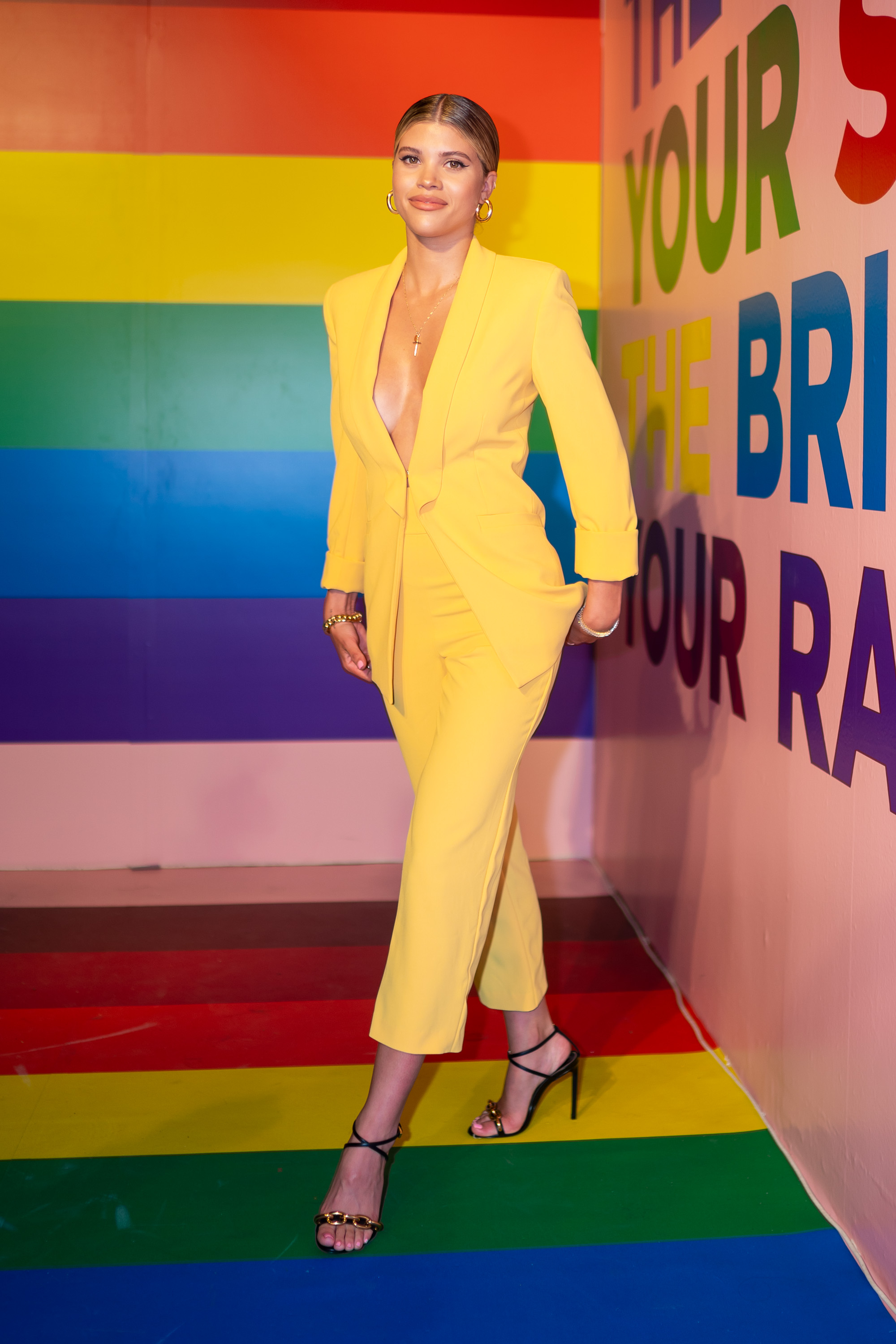 Sofia Richie Poses in a Yellow Suit Jacket With No Shirt and Yellow Pants and Strappy Heels Against a Rainbow Wall