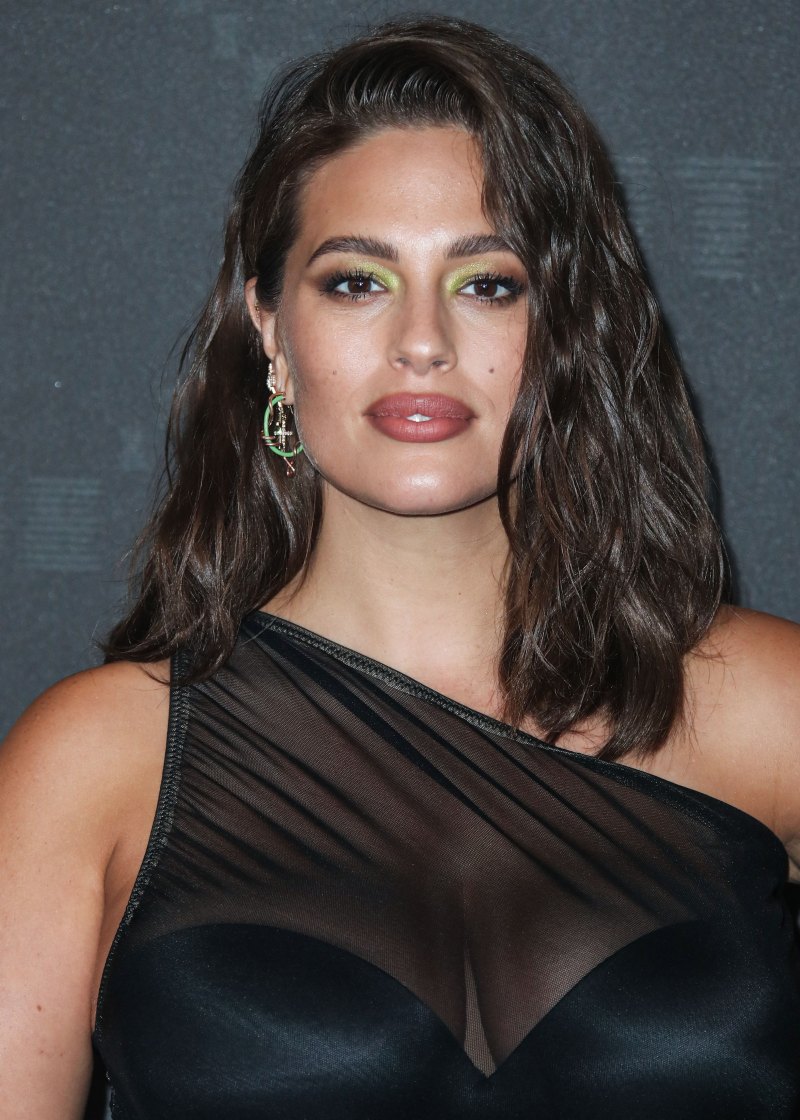 A pregnant Ashley Graham wearing a sheer, black dress and carrying a yellow handbag