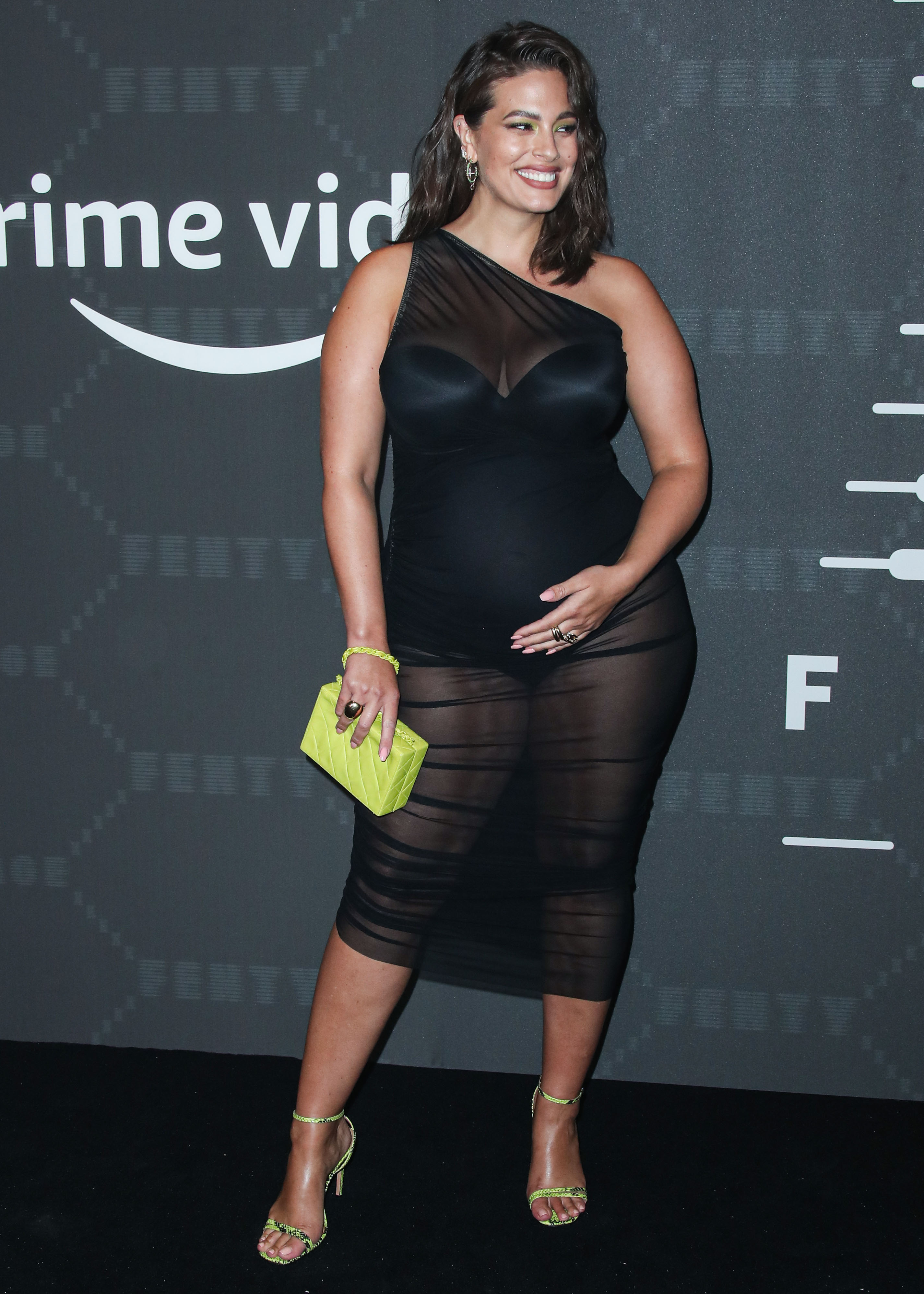 A pregnant Ashley Graham wearing a sheer, black dress and carrying a yellow handbag