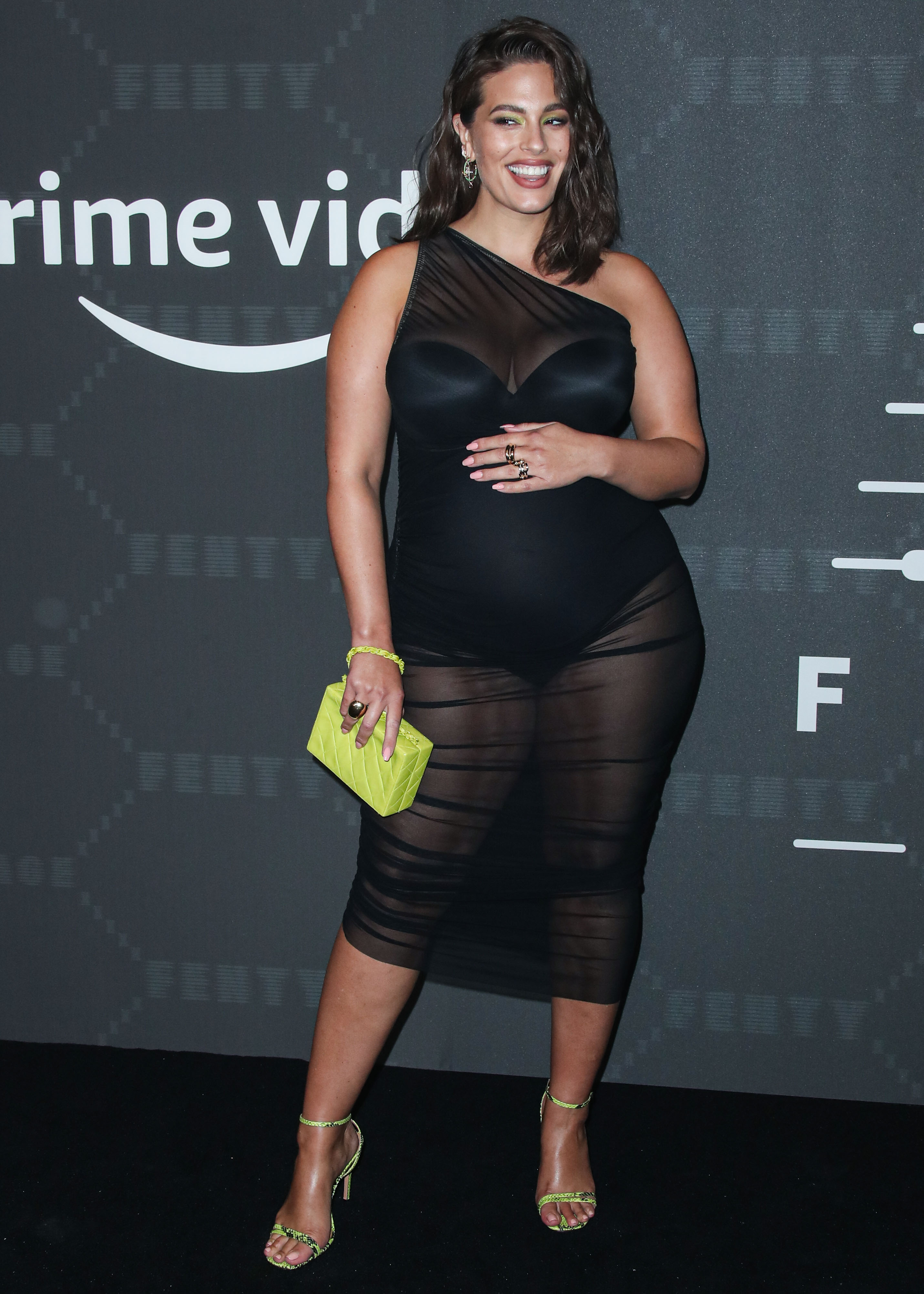 A pregnant Ashley Graham wearing a sheer, black dress and carrying a yellow handbag