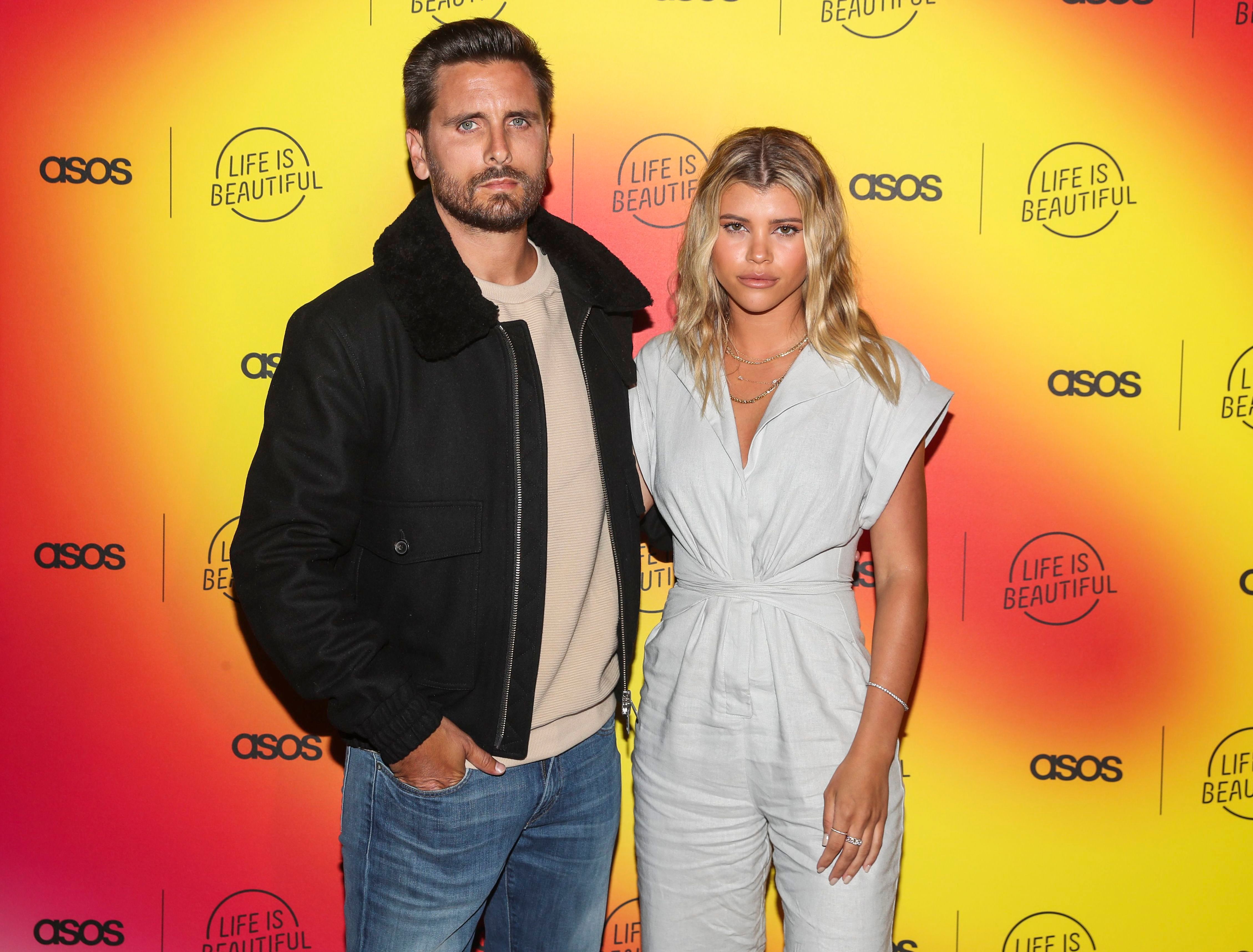 Sofia Richie Wears Grey Jumpsuit Standing With Boyfriend Scott Disick in Jeans and Tan Tshirt and Jacket
