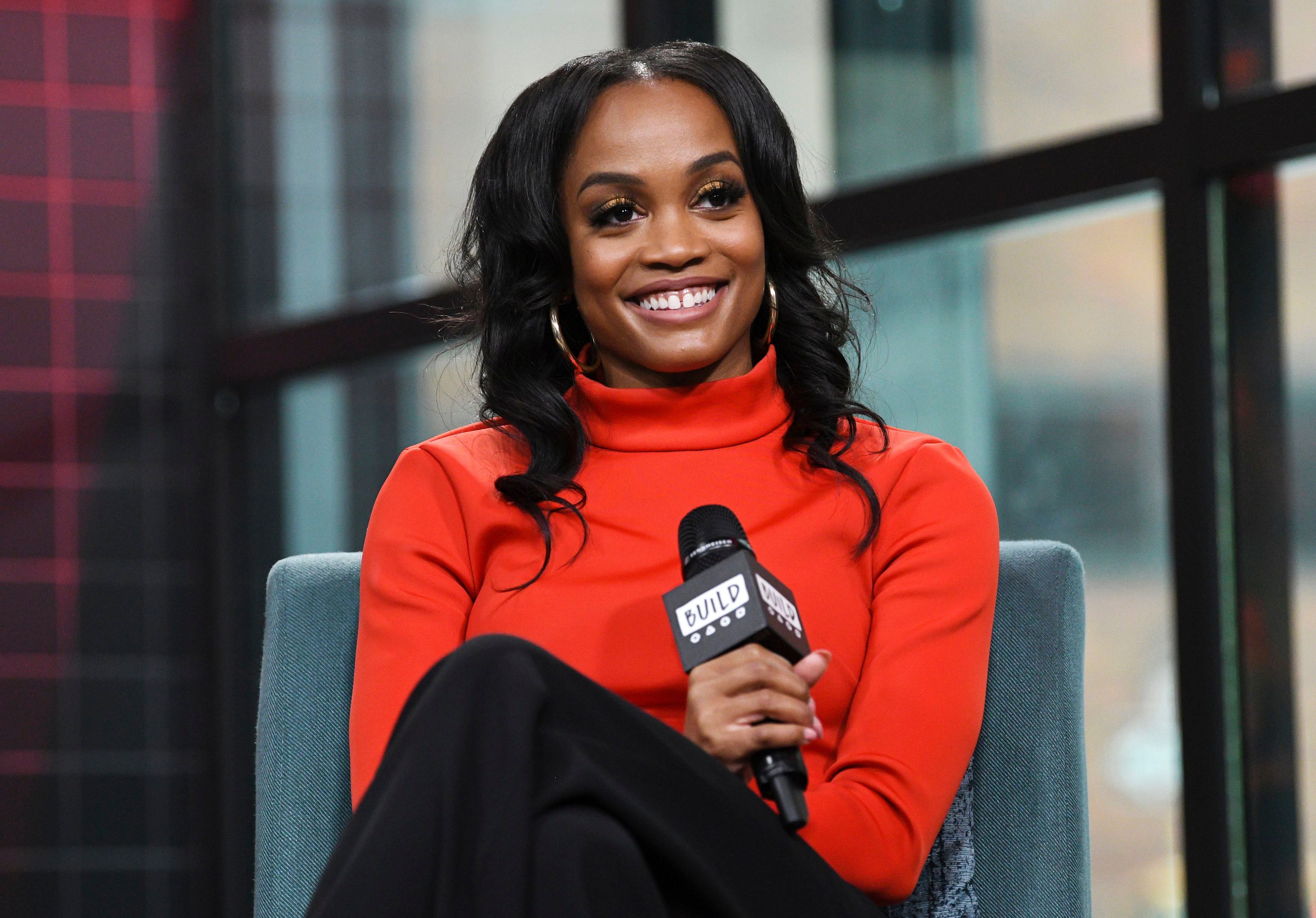Bachelorette Rachel Lindsay Wears Red Turtleneck and Hoop Earrings While Smiling Holding a Microphone