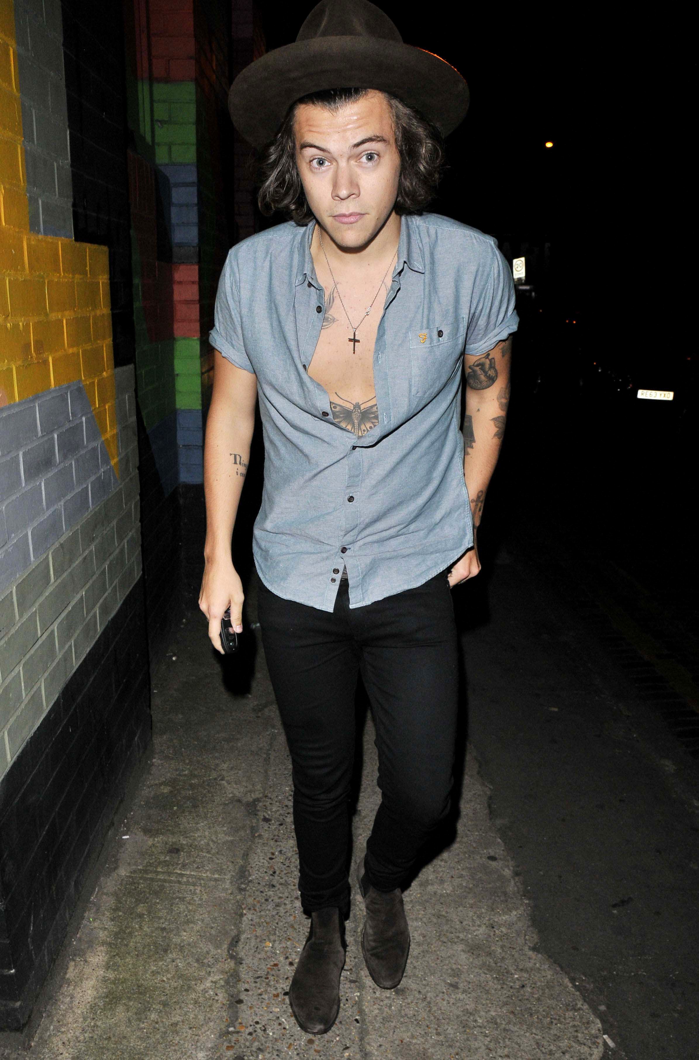 Harry Styles walking outside wearing a blue button-down shirt and a brown hat