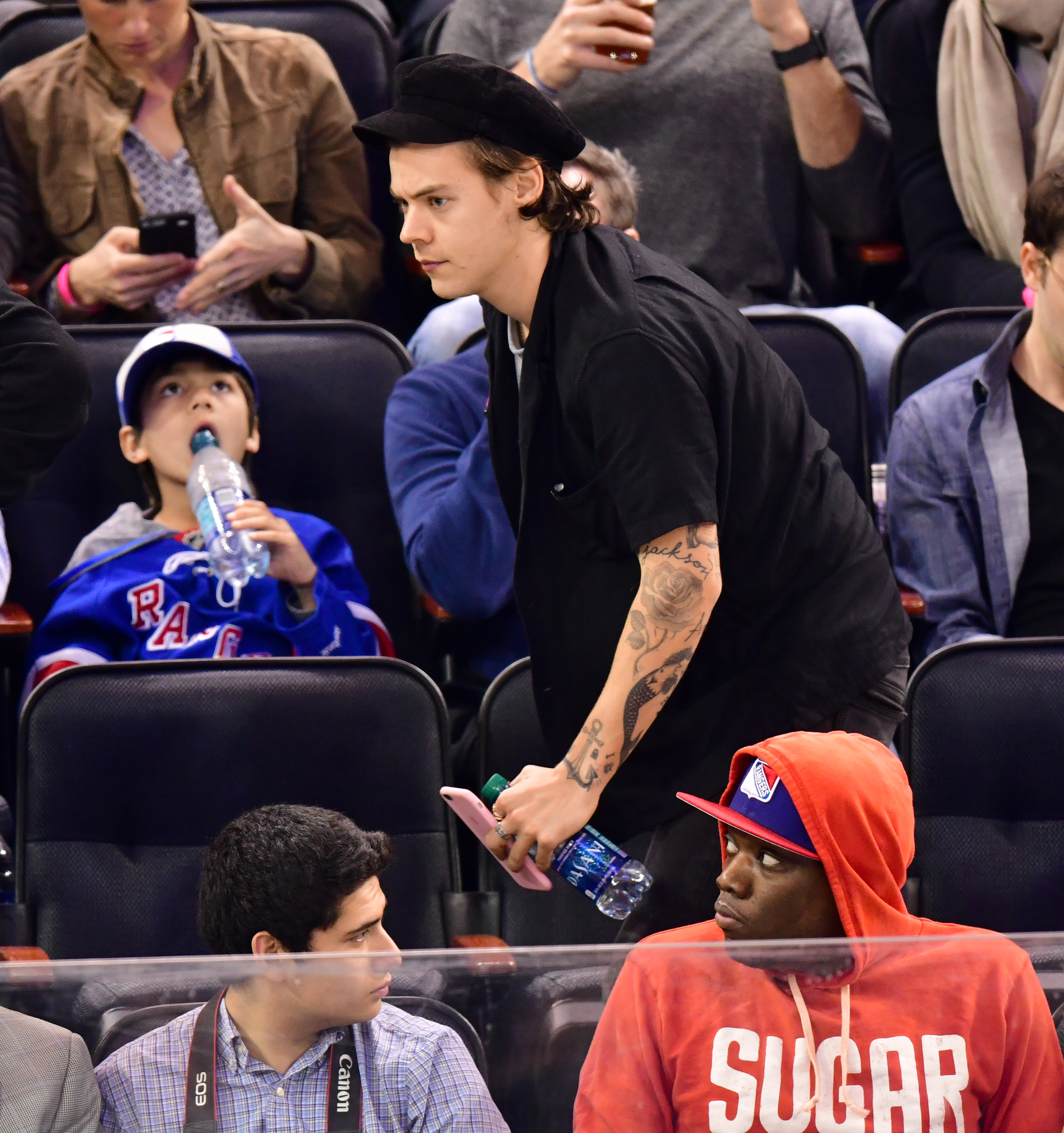 Harry Styles walking to his seat while at a game, wearing a black outfit and hat