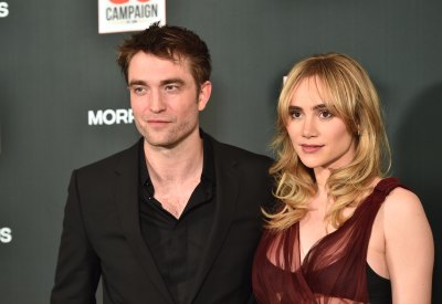 Robert Pattinson, wearing a black suit, stands next to Suki Waterhouse who's wearing a burgundy dress