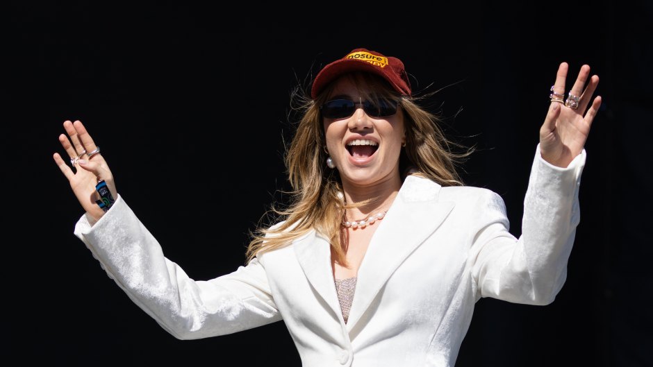 Suki Waterhouse performing on stage while wearing a white jacket, baseball cap and sunglasses