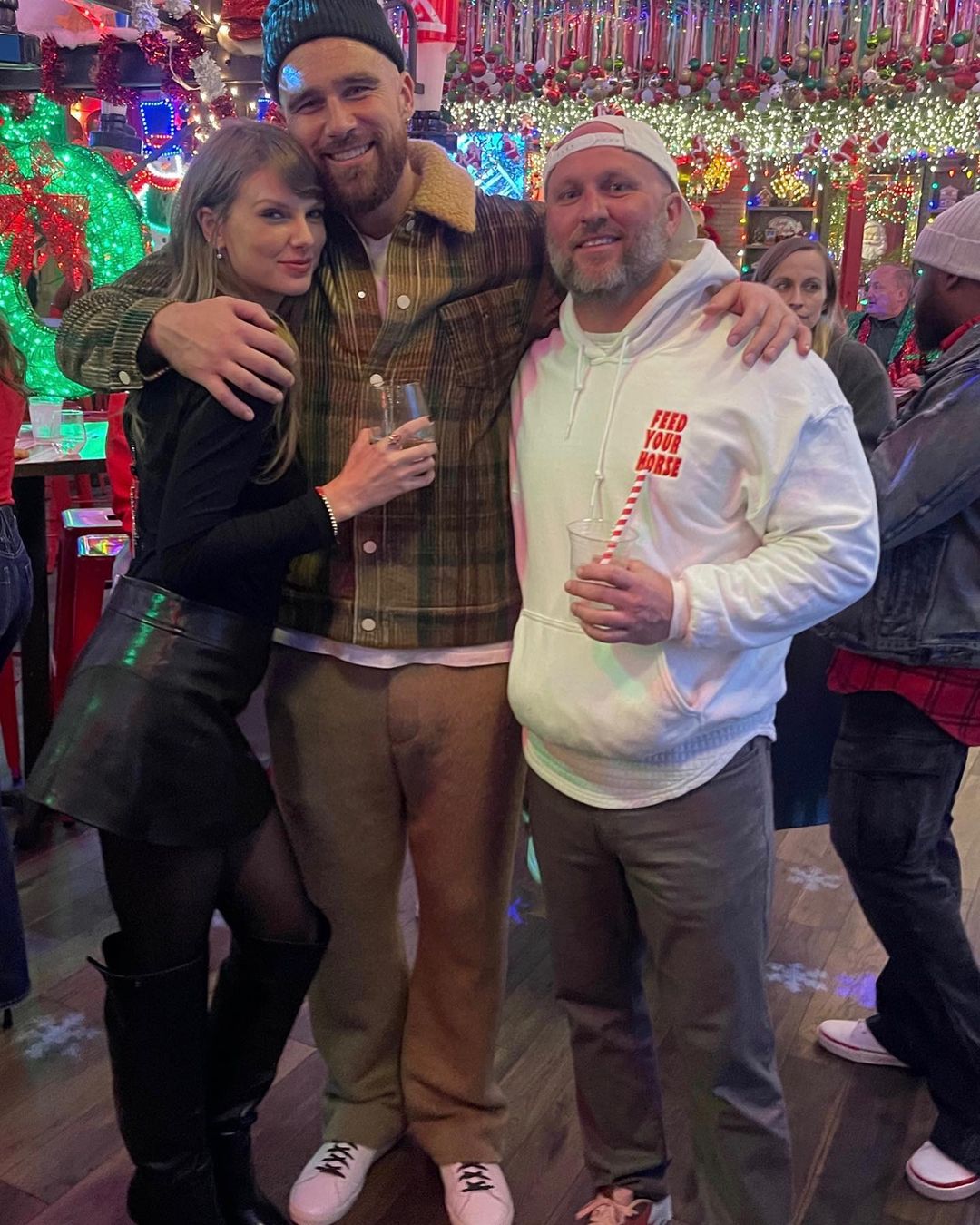 Travis Kelce with his arm around Taylor Swift and a fan during a Christmas party