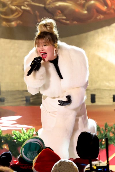 Kelly Clarkson wears a white fur coat while holding a mic on stage.